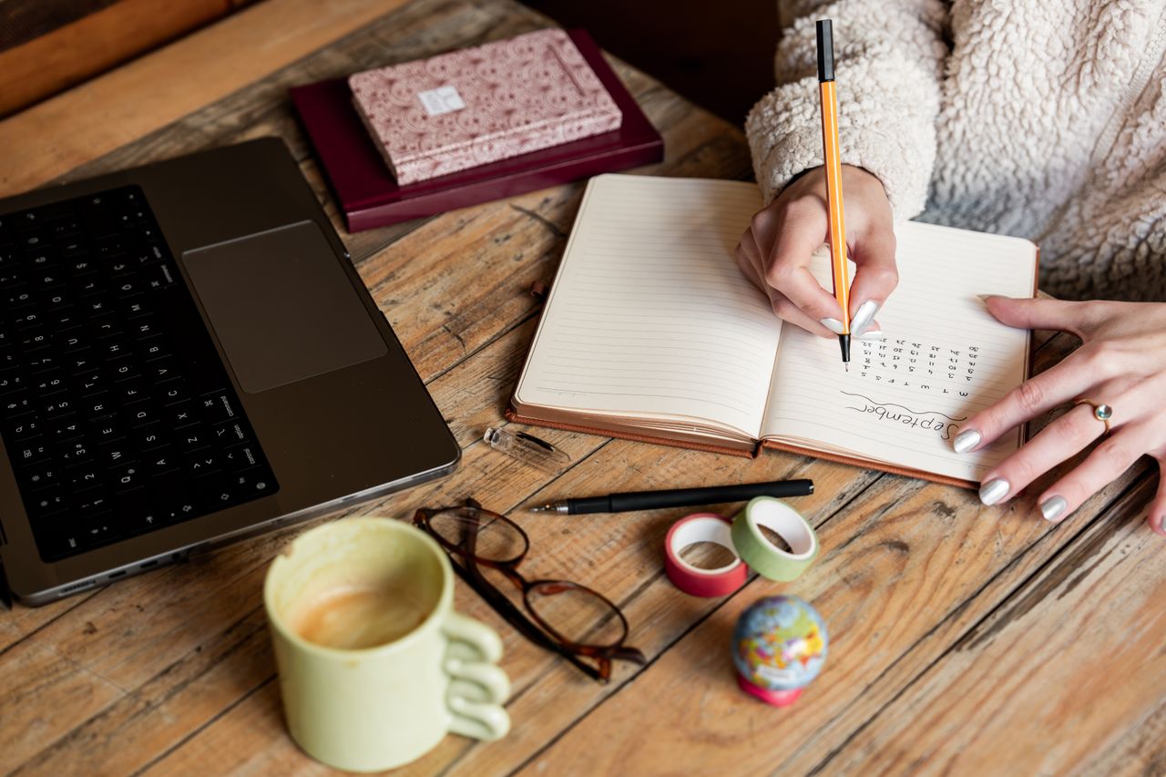 Home A woman planning her schedule in a notepad beside a laptop and coffee on a wooden desk.