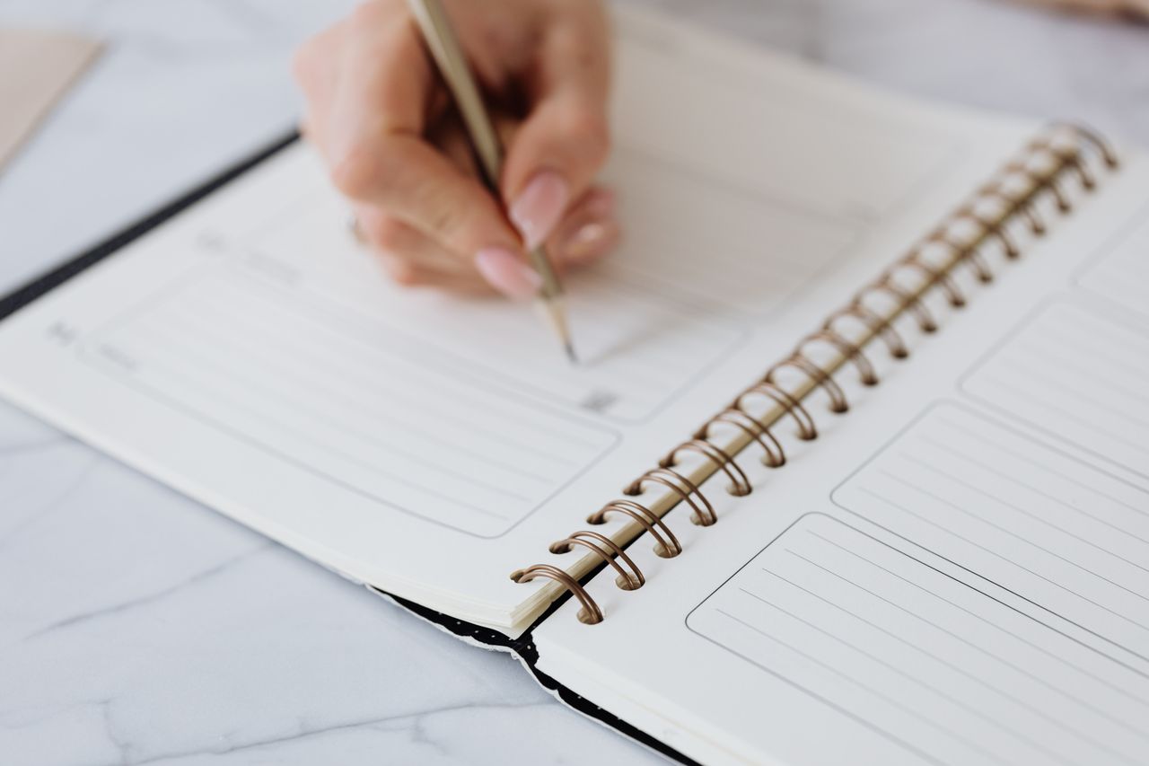 A person writing in a planner with a pencil on a marble surface.