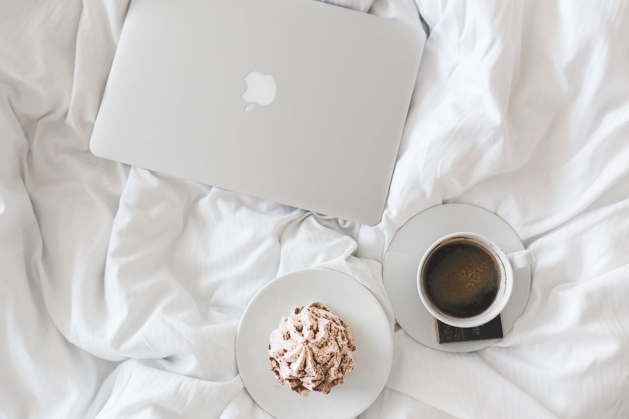 Home A serene morning setup with a laptop, coffee, and cake on a bed, embodying comfort and relaxation.